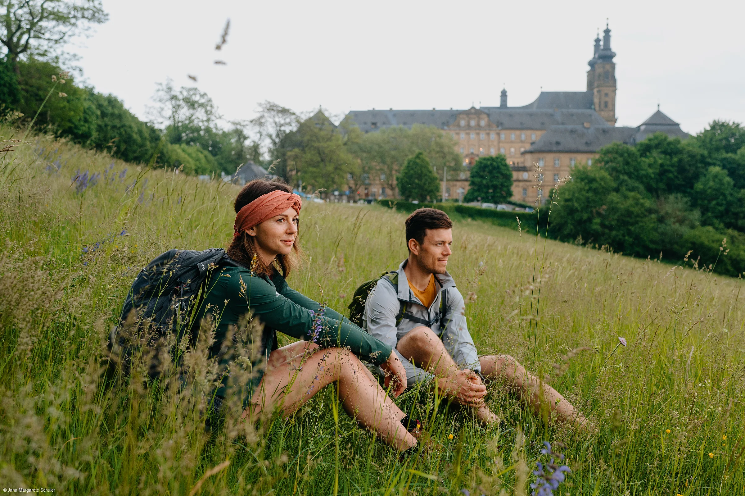Zwei Wandernde machen Rast auf einer Wiese, im Hintergrund eine Klosteranlage mit Türmen