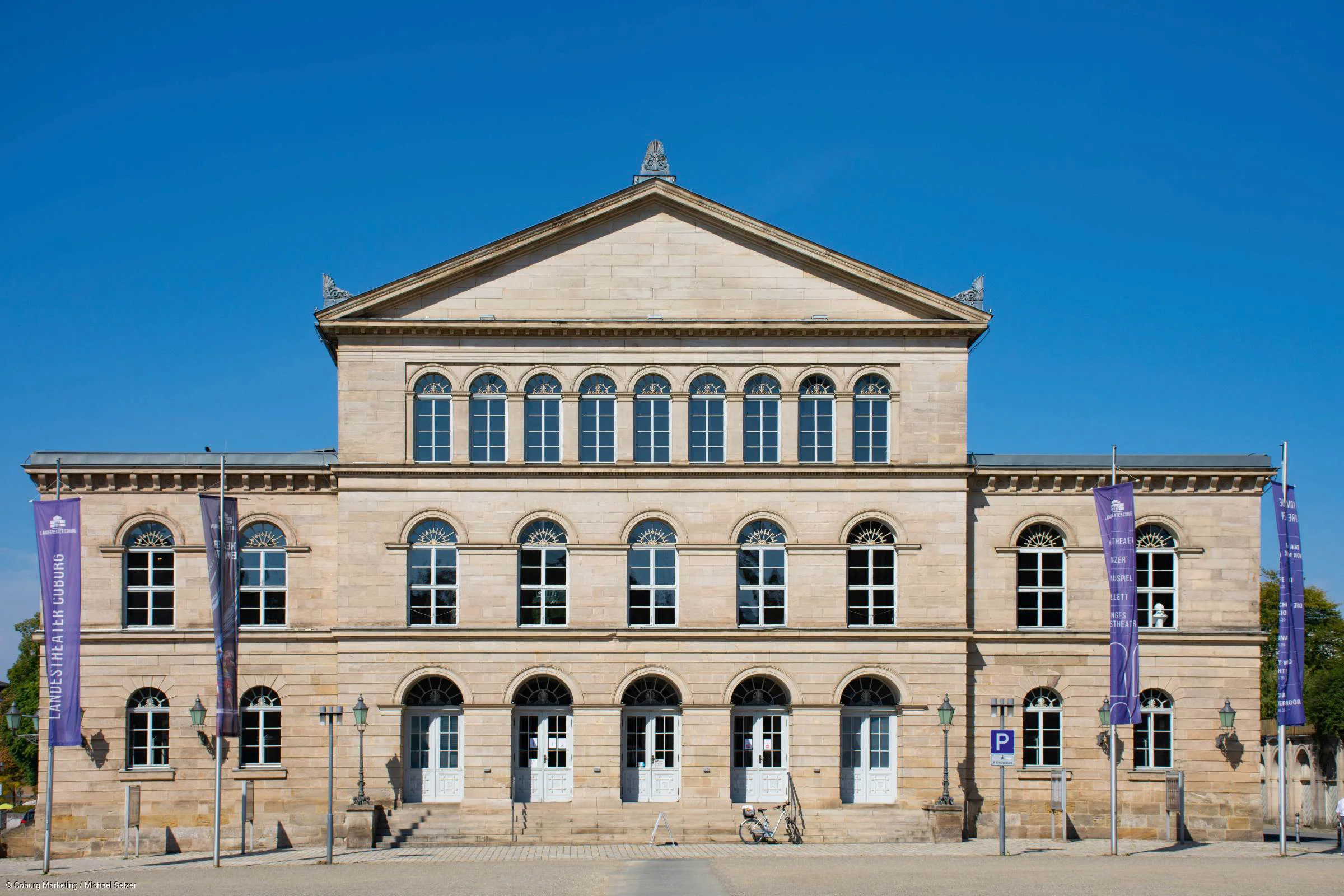 Fassade eines historischen Gebäudes mit vielen Fenstern und vier weißen Türen bei klarem Himmel.