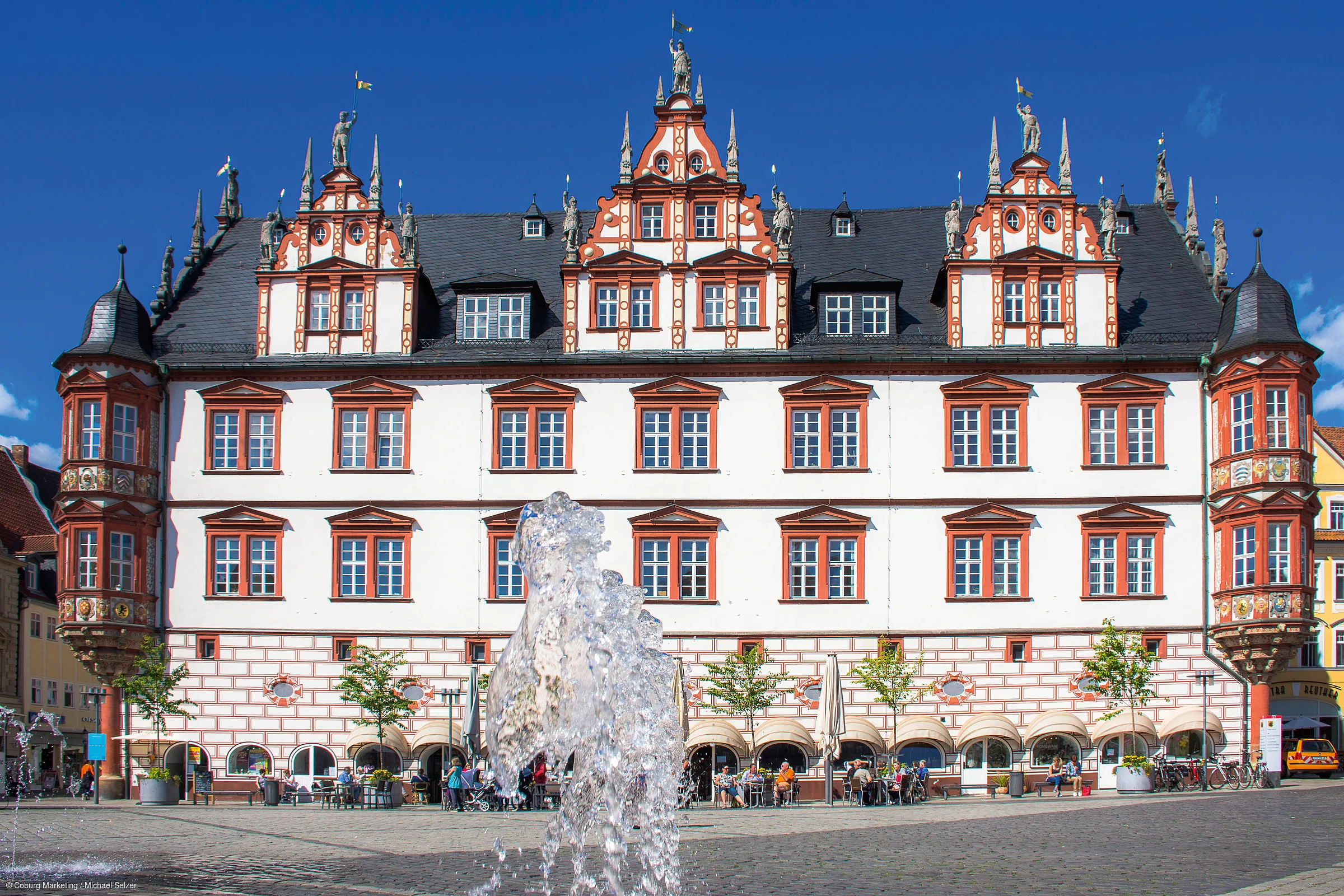 Historisches Gebäude mit roten Fensterrahmen und Wasserspielen auf dem Platz davor bei blauem Himmel.